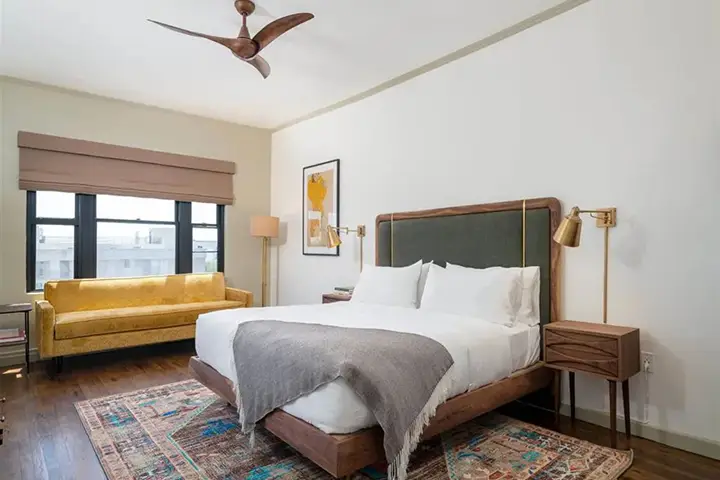 Modern bedroom with wooden bed, white bedding, yellow sofa, and ceiling fan.