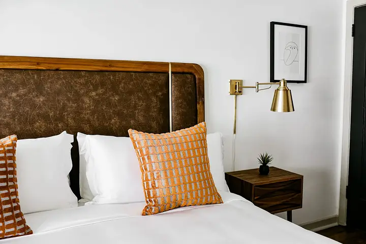 Neatly made bed with orange pillows, wall lamp, and line art on white wall.