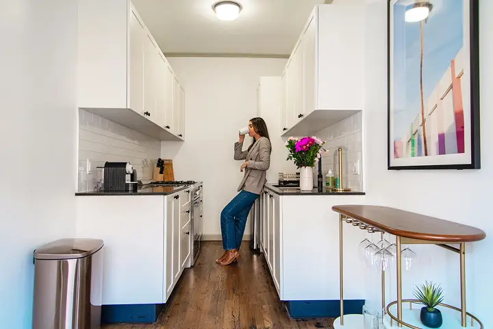 Woman sipping cup in white kitchen with flowers and stainless appliances.
