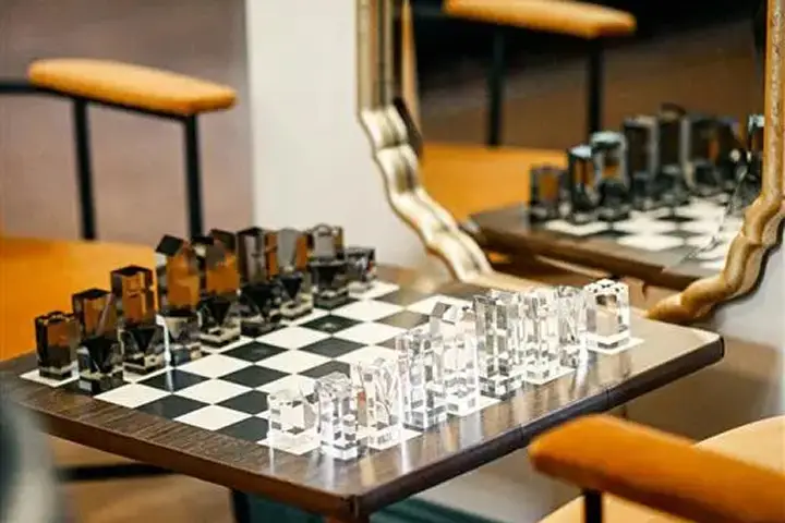 Glass chess set on wooden board with orange chairs in the background.