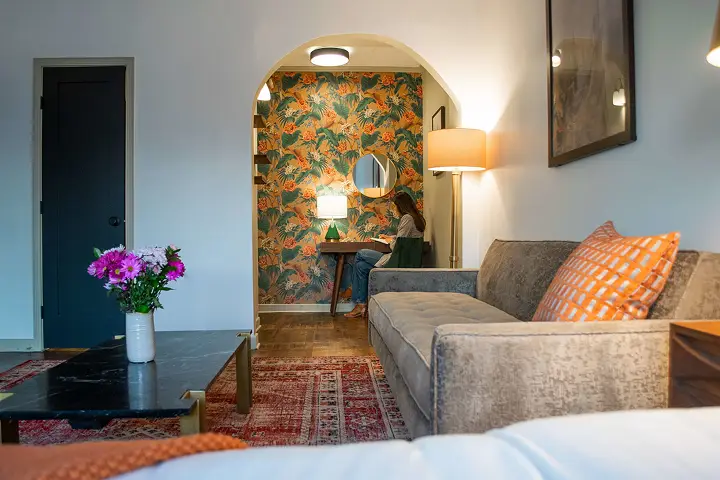 Reading nook with floral wallpaper, cozy sofa, colorful cushions, and flowers.