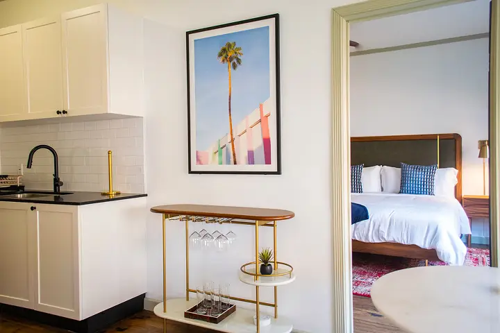 Small kitchen, bar cart, and cozy bedroom with framed palm tree photo.