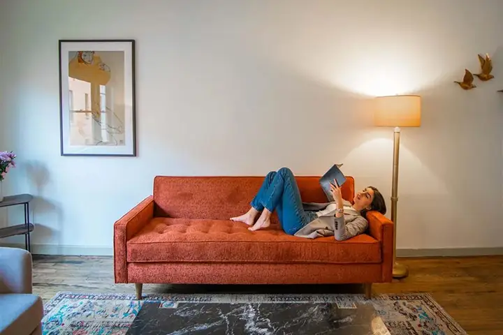 Person reading on orange sofa with floor lamp, flowers, and wall art.